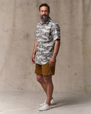 Man wearing a camouflage shirt and brown shorts standing against a concrete wall.