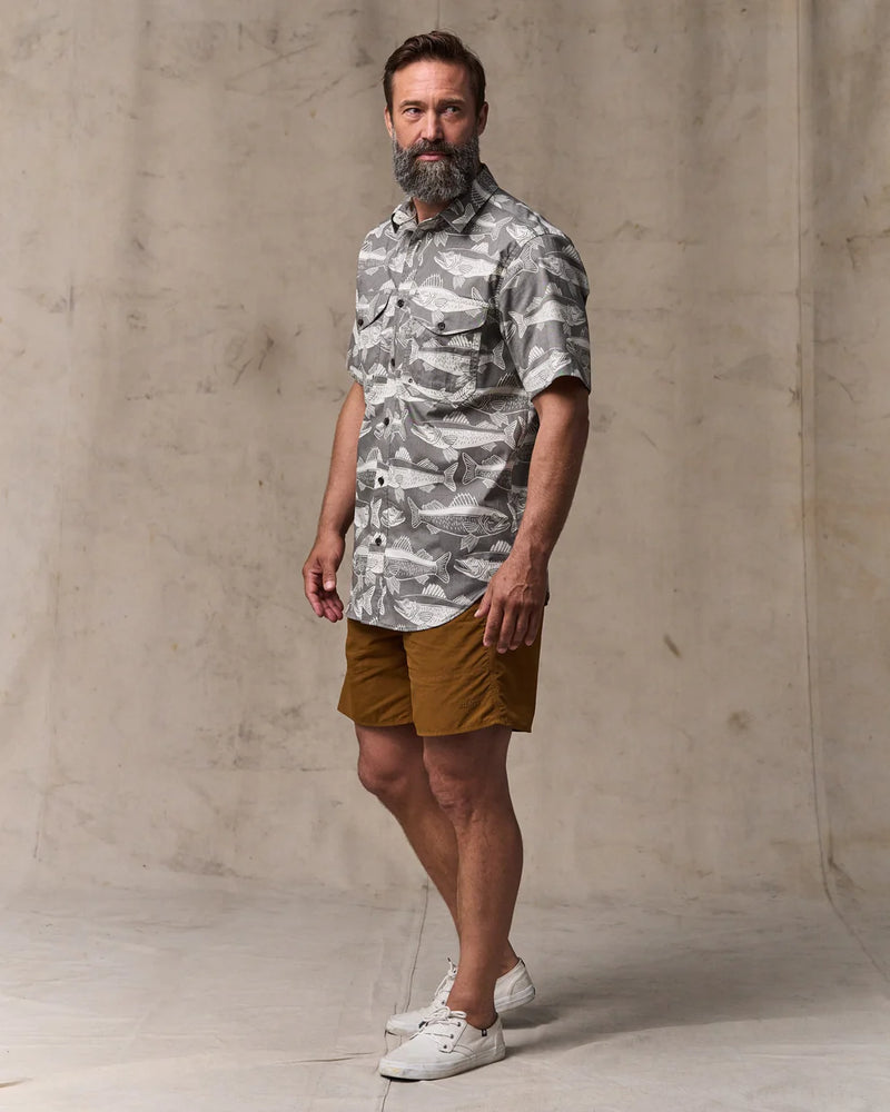 Man wearing a camouflage shirt and brown shorts standing against a concrete wall.
