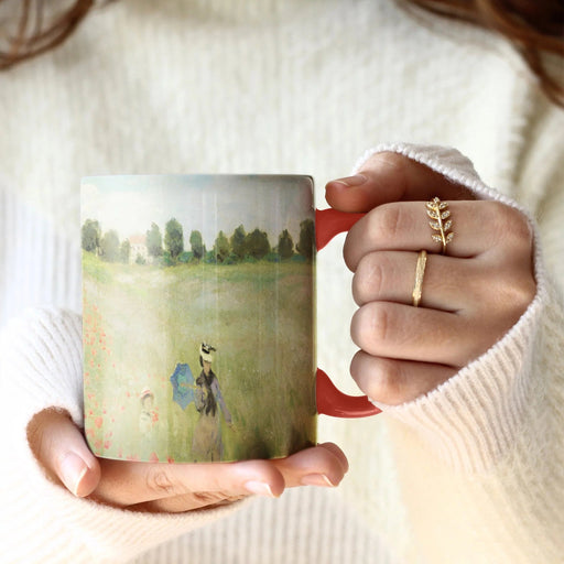 Woman with shoulder-grazing brown hair in a cozy cream sweater wearing two golden rings, one a slim band and the other a vine with leaves and pavé, holds a mug with both hands - one by the handle, the other hand supporting the bottom of the mug. The mug features a full-coverage print of Monet's Poppy Field painting and a red handle.