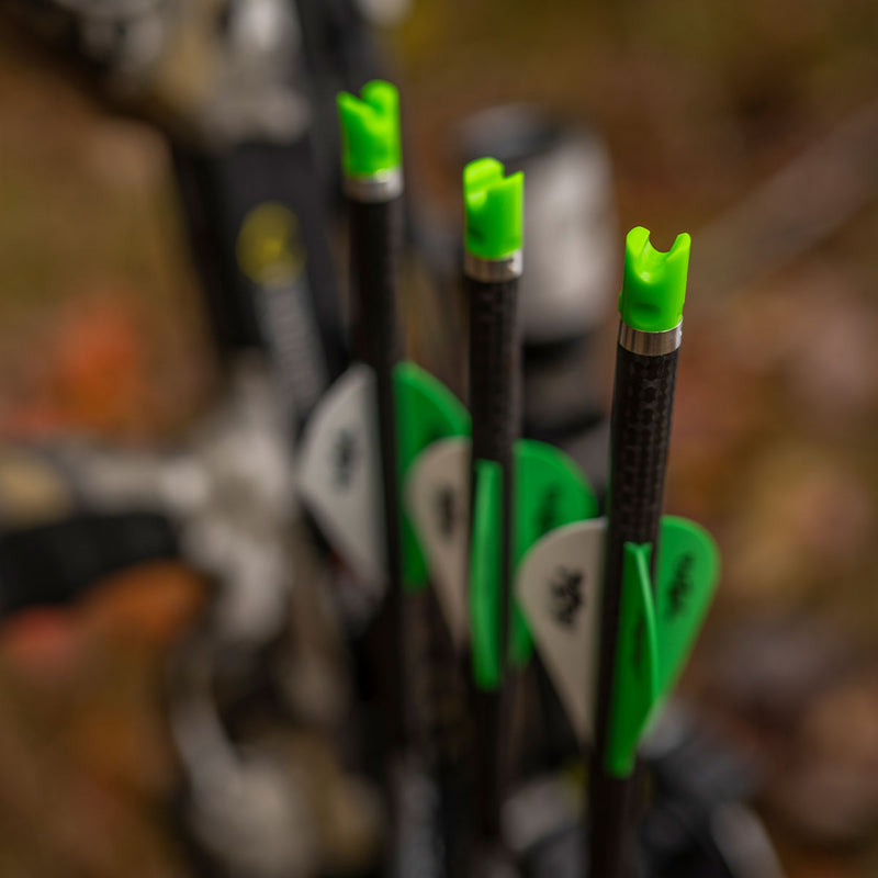 Close-up of several arrows with bright green nocks and green-and-white fletching, standing upright in a blurred outdoor setting.