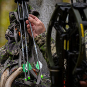 Person in camouflage holding a bow with arrows, surrounded by nature.