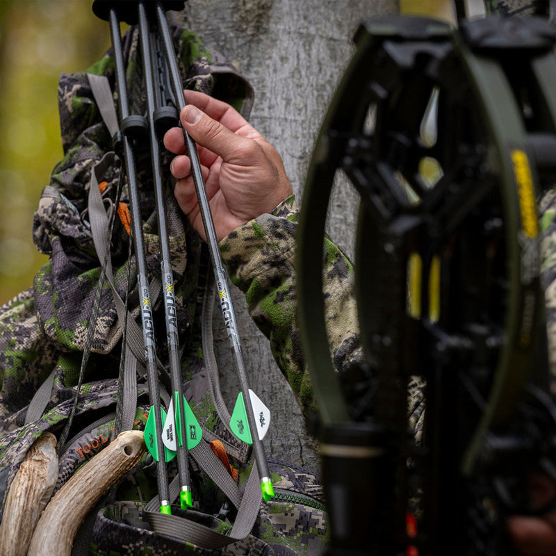 Person in camouflage holding a bow with arrows, surrounded by nature.