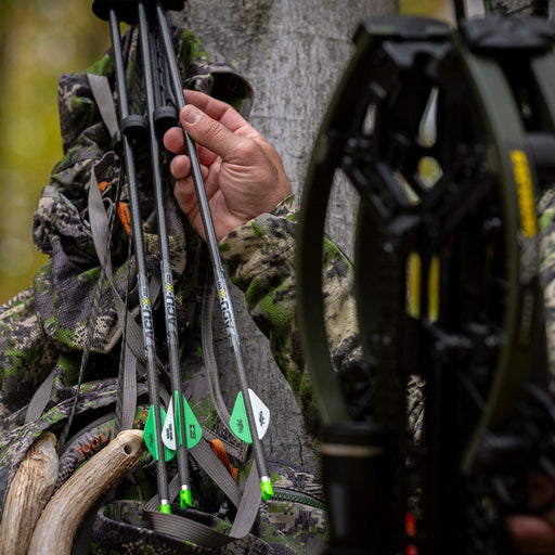 Person in camouflage holding a bow with arrows, surrounded by nature.