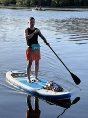Man wearing a blue waterproof belt bag standing on a white paddleboard with blue and black accents in a lake. 