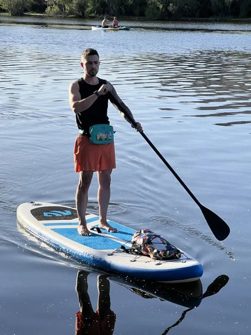 Man wearing a blue waterproof belt bag standing on a white paddleboard with blue and black accents in a lake. 