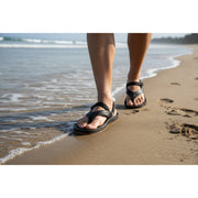 A man walking down the coastline wearing a pair of black sandals.