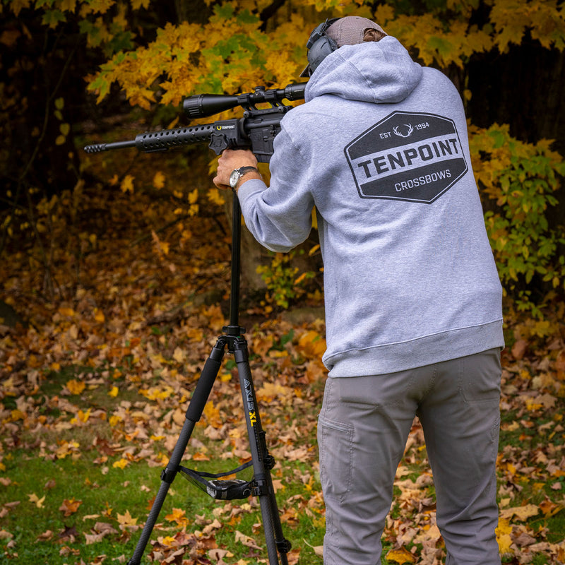 Person wearing a Tenpoint hoodie with a hunting rifle on a tripod in an outdoor setting.