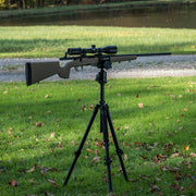 A rifle with a mounted scope sits on a black tripod set on a grassy lawn with scattered leaves, outdoors near a path and water.