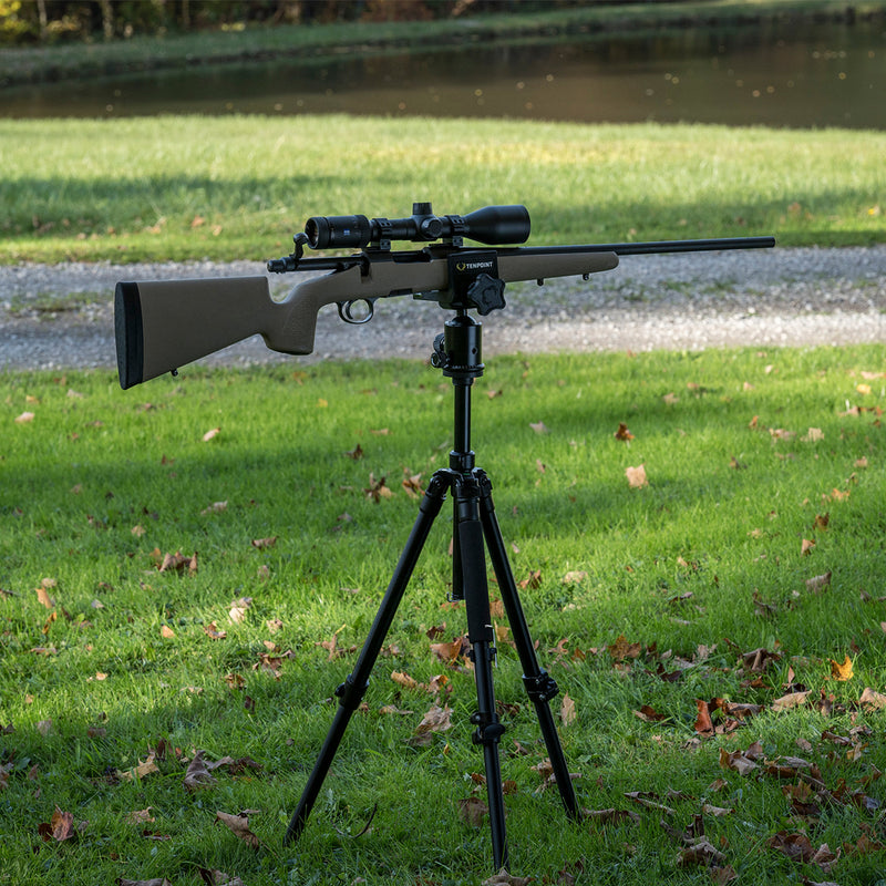 A rifle with a mounted scope sits on a black tripod set on a grassy lawn with scattered leaves, outdoors near a path and water.
