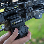 A close-up of a person's hands adjusting a black Tenpoint accessory mounted on a black and camouflage bow, with a blurred green outdoor background.