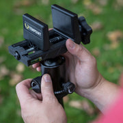 A close-up of a person's hands adjusting a black TenPoint tripod head clamp outdoors on a grassy background.