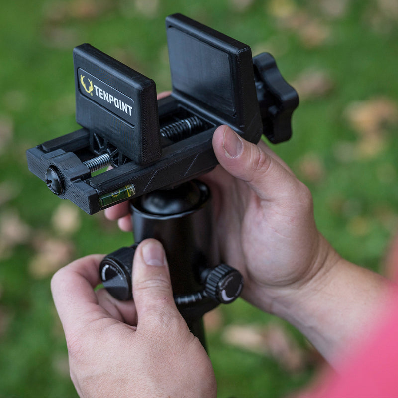 A close-up of a person's hands adjusting a black TenPoint tripod head clamp outdoors on a grassy background.