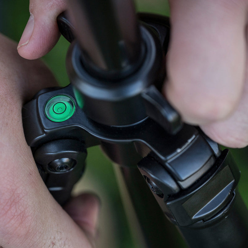 Close-up of a person's hands adjusting the locking mechanism of a black tripod, with a bright green bubble level visible on the side.