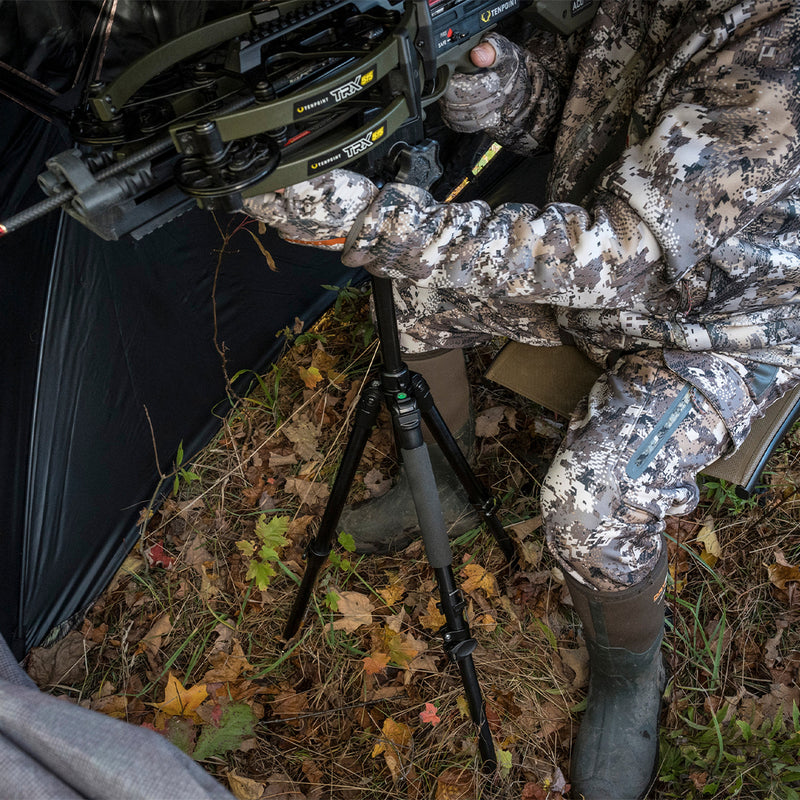 Hunter in camouflage gear with a bow and tripod in a forest setting