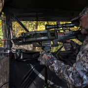 A man in camouflage clothing aims a TenPoint crossbow from inside a hunting blind, with trees visible in the background.