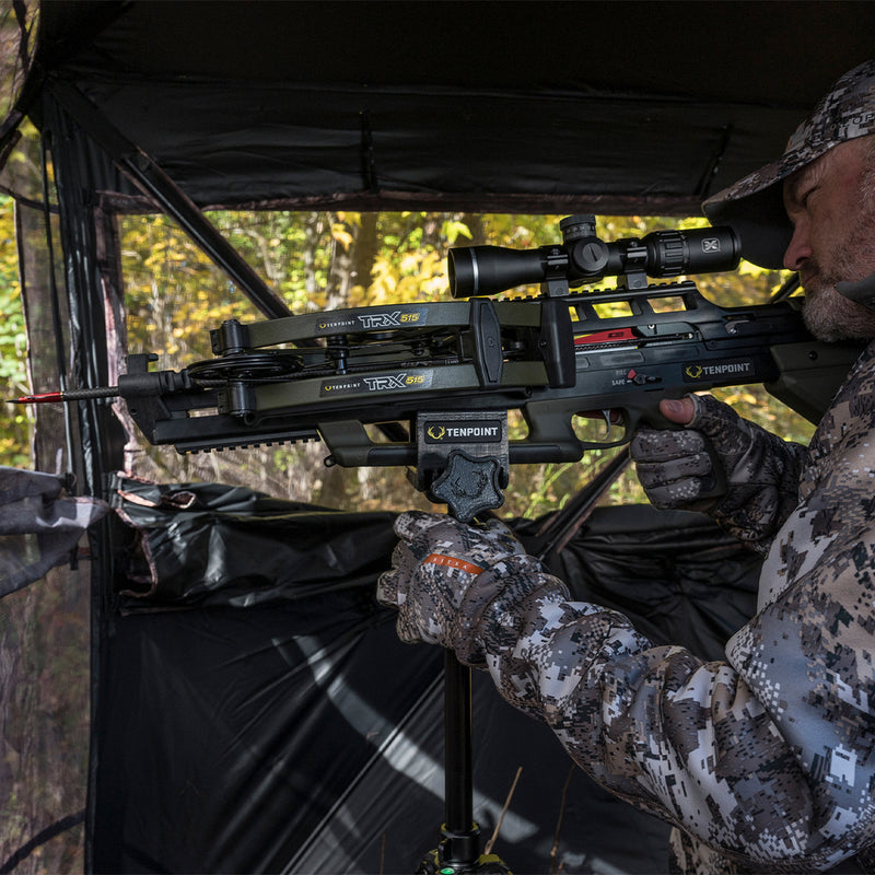 A man in camouflage clothing aims a TenPoint crossbow from inside a hunting blind, with trees visible in the background.