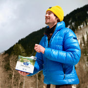 Person in blue jacket and yellow beanie holding a Nature Valley bar outdoors with mountains in the background