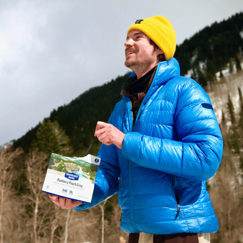 Person in blue jacket and yellow beanie holding a Nature Valley bar outdoors with mountains in the background