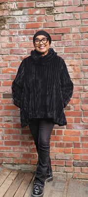 Person wearing a black poncho and glasses standing against a brick wall.
