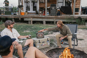 Two men sitting by a fire pit with a cooler, surrounded by outdoor equipment.