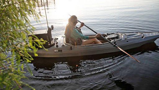 Woman paddling her grey fishing kayak through calm waters near a tree.