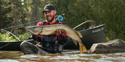 A man standing in a river holding up a giant fish with a black canoe and fishing gear in the background. The canoe has a Rangeley 15 logo on the side of it.