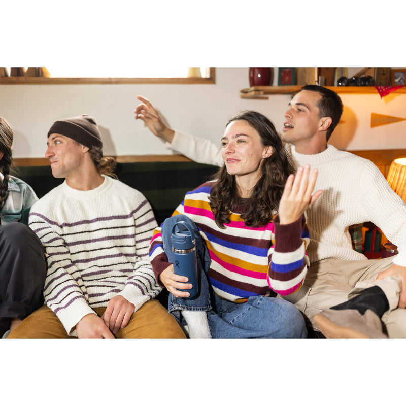 Four young adults sitting together in a cozy living room setting, wearing casual knit sweaters and jeans. The person in the center holds a blue insulated tumbler with a white lid.