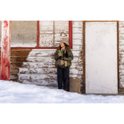 A woman wearing an olive green hooded jacket, dark pants, and a beige beanie stands outdoors in front of a weathered white wooden building with snow in the foreground. She is holding an olive green travel mug.