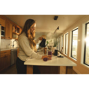 A woman in a modern kitchen pours hot water from a black kettle into a coffee maker on a white countertop. The scene is warmly lit by sunlight coming through large windows, with wooden cabinets and kitchen appliances visible in the background.