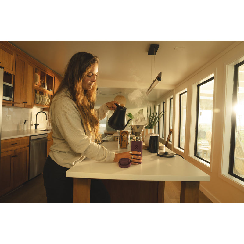 A woman in a modern kitchen pours hot water from a black kettle into a coffee maker on a white countertop. The scene is warmly lit by sunlight coming through large windows, with wooden cabinets and kitchen appliances visible in the background.
