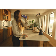 A woman in a modern kitchen setting drinking from a blue insulated tumbler cup with a metal lid, standing beside a kitchen island.