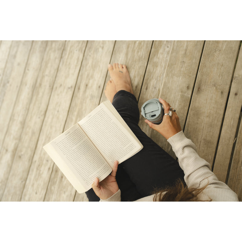 A top-down view of a person sitting on a wooden deck, holding an open book and a gray travel tumbler cup.