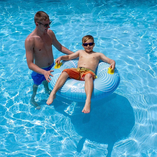 A child floating in a swimming pool using a blue and yellow inflatable tube while his father helps him.