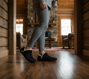 Person wearing black slippers in a cozy living room with wooden floors and furniture.