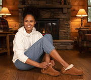 Woman sitting on the floor wearing brown suede shoes with white fur trim on the floor of a cozy cabin