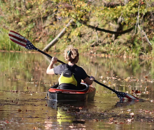 Back view of a woman paddling a red kayak down a river with an americana patterned paddle.