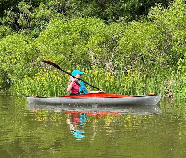 Woman paddling her red kayak down a river next to some tall grass.