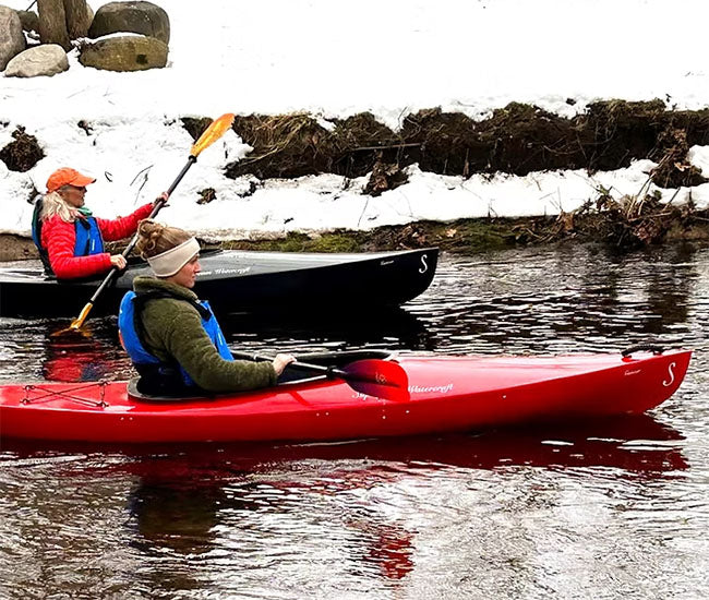 Two women paddling their kayaks down a calm river on a snowy background.
