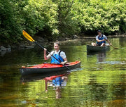 A woman in a red kayak paddling down a river with a man in a green kayak behind her.