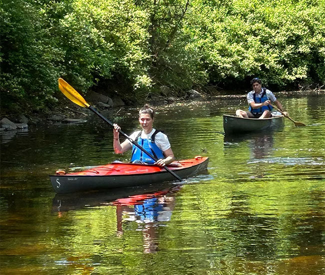 A woman in a red kayak paddling down a river with a man in a green kayak behind her.
