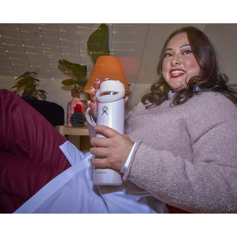 A woman in a cozy indoor setting holds a white insulated water bottle with a matching lid. She is wearing a sparkly light pink sweater and white pants, with a small table, plants, and warm string lights in the background.