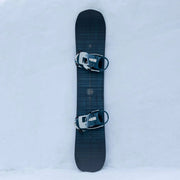 Top view of a black snowboard with bindings attached against a snowy hill. 