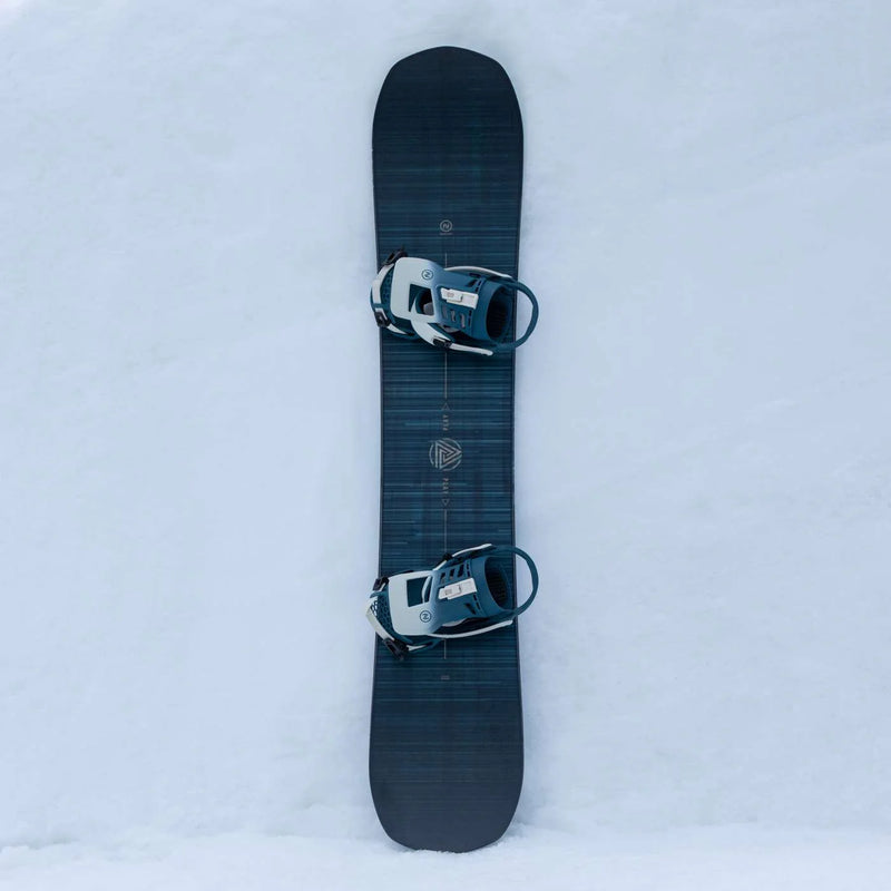Top view of a black snowboard with bindings attached against a snowy hill. 