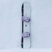 White snowboard with black accents and pink bindings on a snowy hill. 