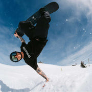 Man doing a handstand riding a black colored snowboard down a snowy mountain. 