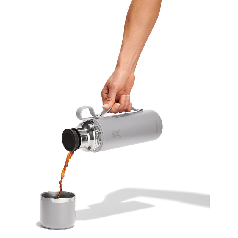 A hand holds a silver insulated bottle with a black lid, pouring dark coffee into a matching silver cup against a clean white background.