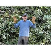 A man wearing a blue long-sleeve shirt, brown work gloves, and a dark cap stands in front of a dense background of green leaves and branches, with both hands raised in a gesture.