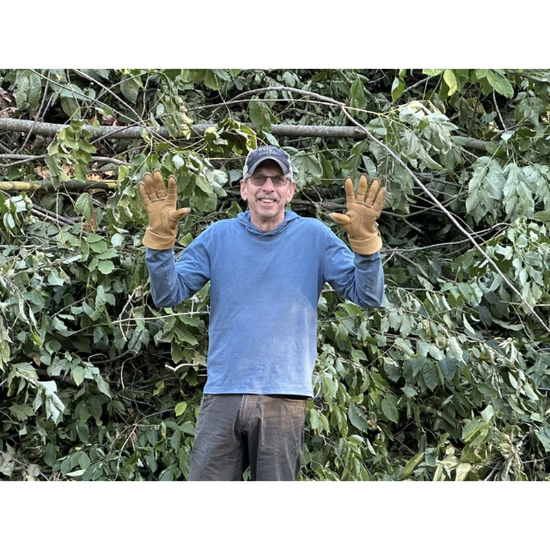A man wearing a blue long-sleeve shirt, brown work gloves, and a dark cap stands in front of a dense background of green leaves and branches, with both hands raised in a gesture.