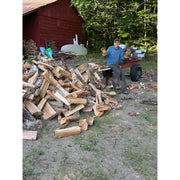 A man leans on a log splitter beside a pile of chopped firewood logs next to a red wooden shed and trees, showing off his Vermonter gloves, holding up perfectly to the use.