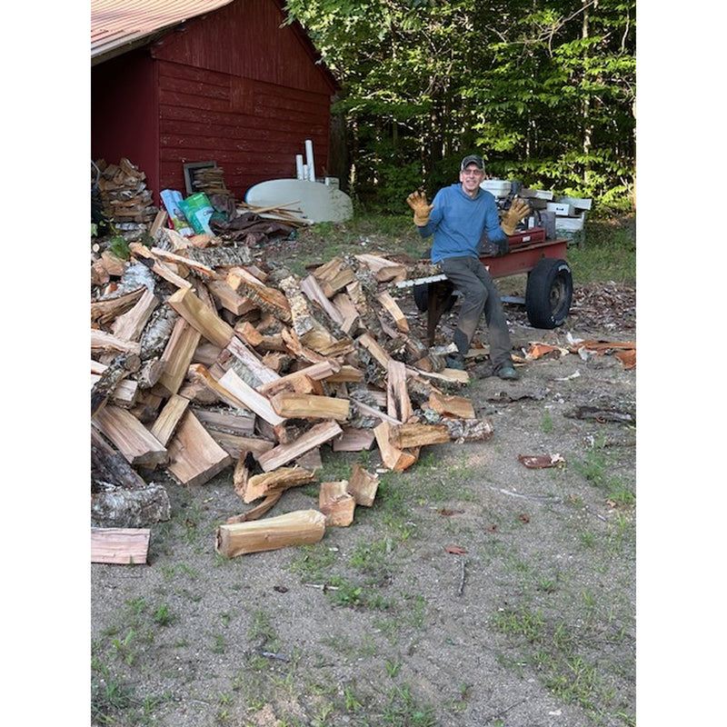 A man leans on a log splitter beside a pile of chopped firewood logs next to a red wooden shed and trees, showing off his Vermonter gloves, holding up perfectly to the use.